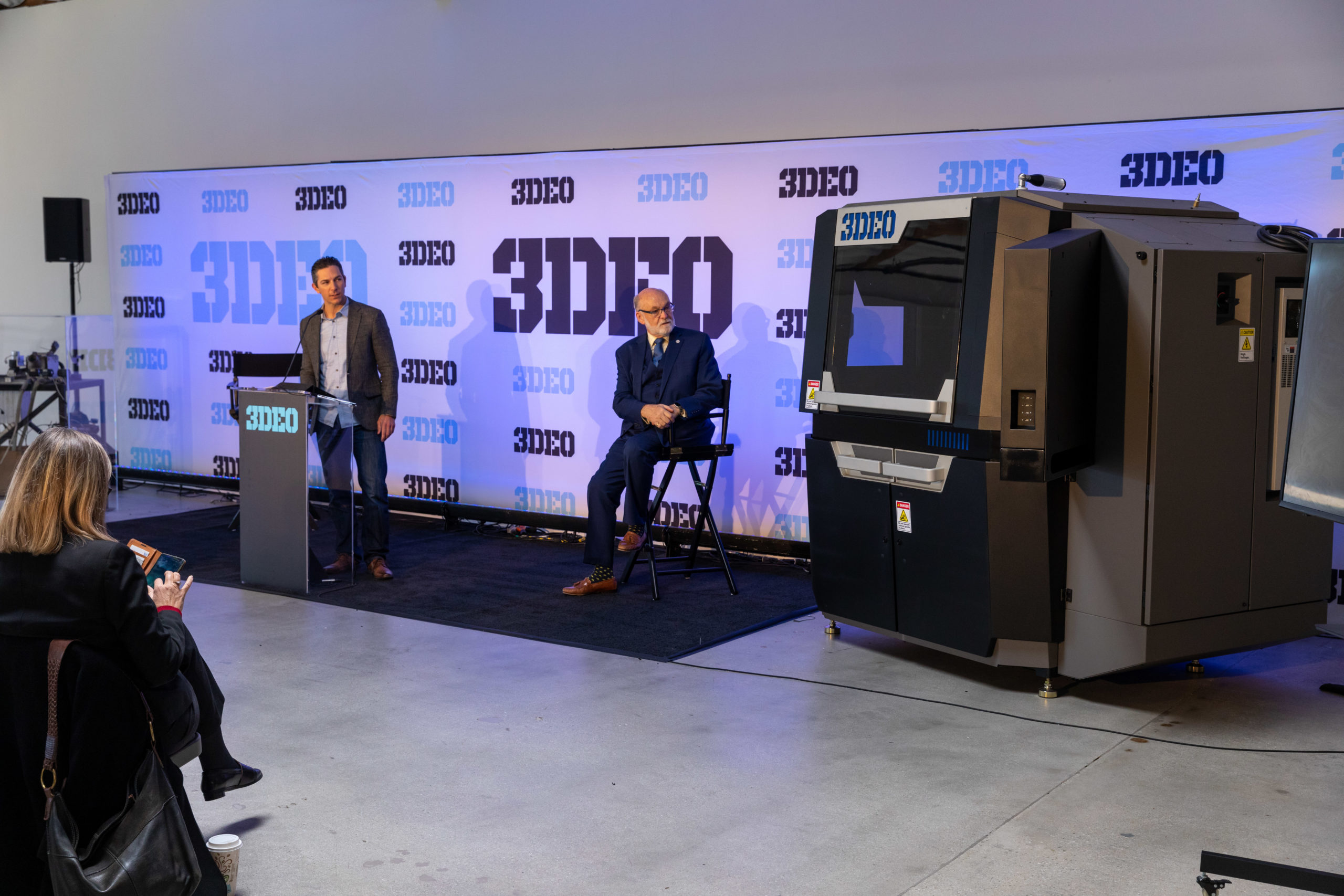 Two men, one standing and one seated, at a presentation in a room with large 3d printers and a branded backdrop with multiple logos.