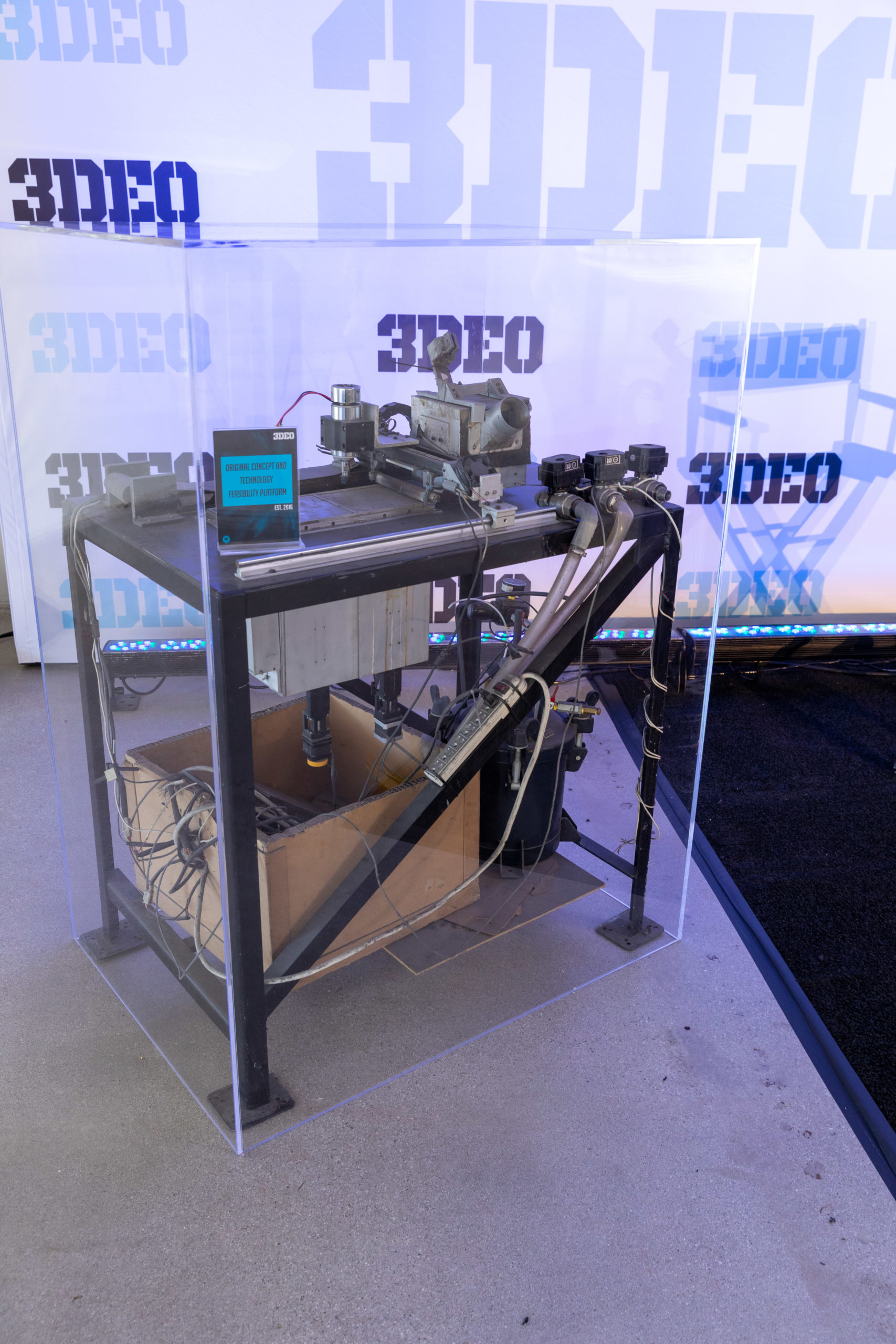 Industrial equipment on display with visible wiring and pipes inside a glass case at a technology expo, with repetitive "3deo" branding in the background.