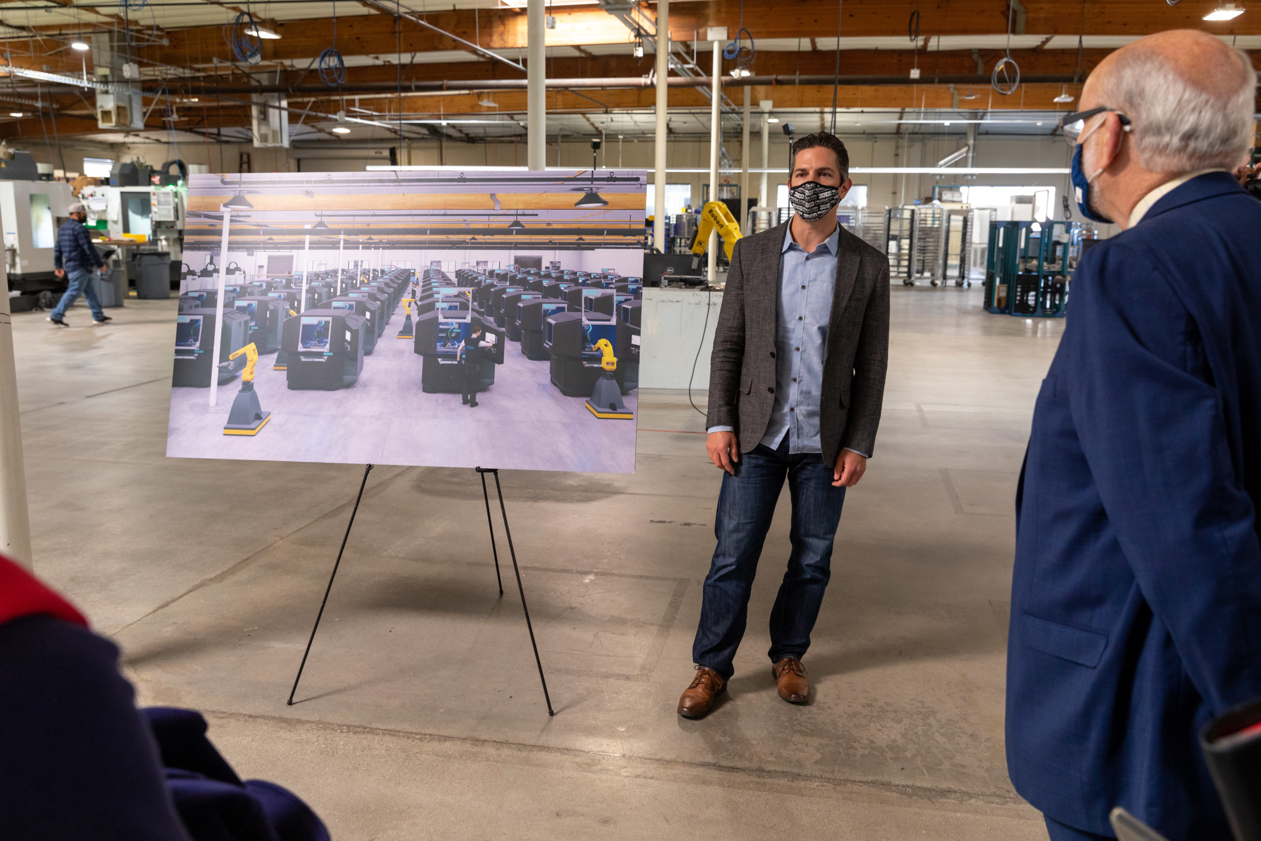 A man in a mask and blazer discusses a large printed photo of a factory floor with another person in a factory setting.