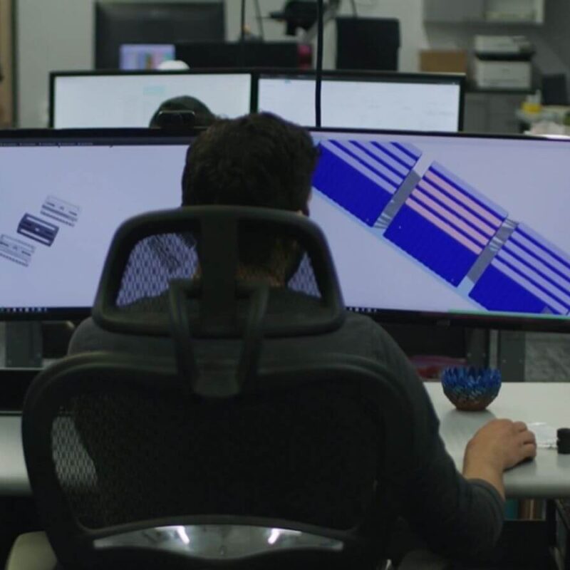 A person sitting at a desk with a large monitor displaying complex diagrams, surrounded by office equipment and other desks in an open-plan office.
