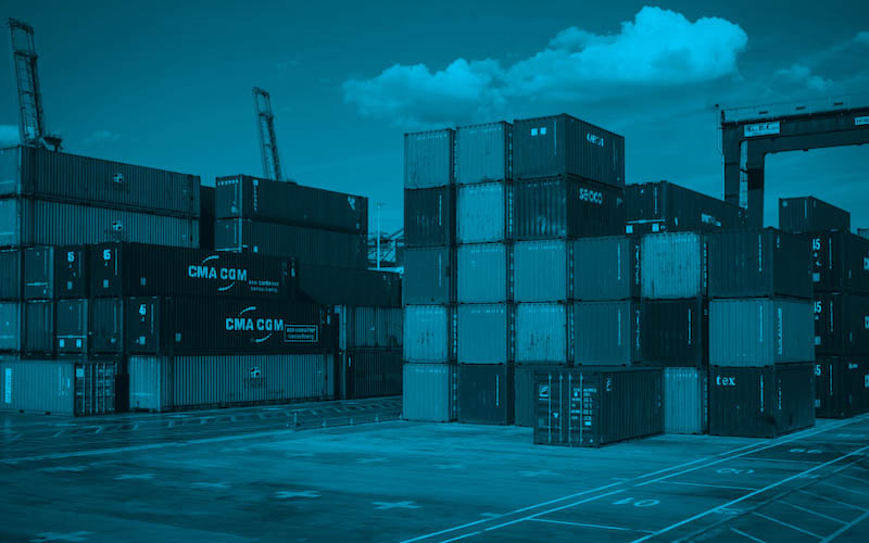 A maritime shipping yard with stacked cargo containers and cranes in the background under a partly cloudy sky.