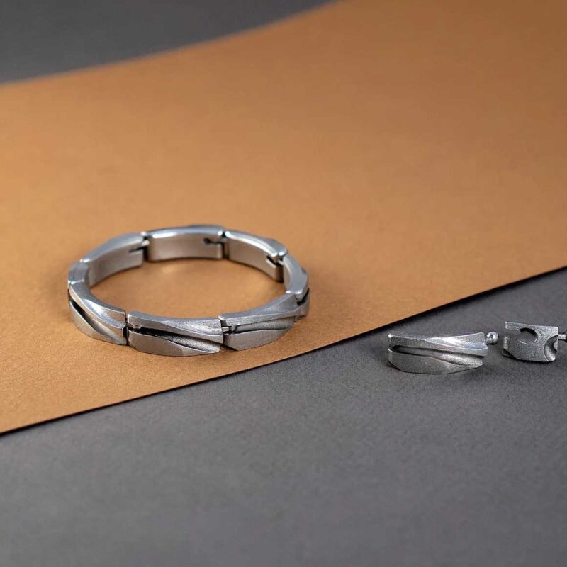 A silver bracelet and a pair of silver earrings, crafted through metal additive manufacturing, are placed on a brown and gray surface. The bracelet features a segmented design, while the earrings have a matching pattern.