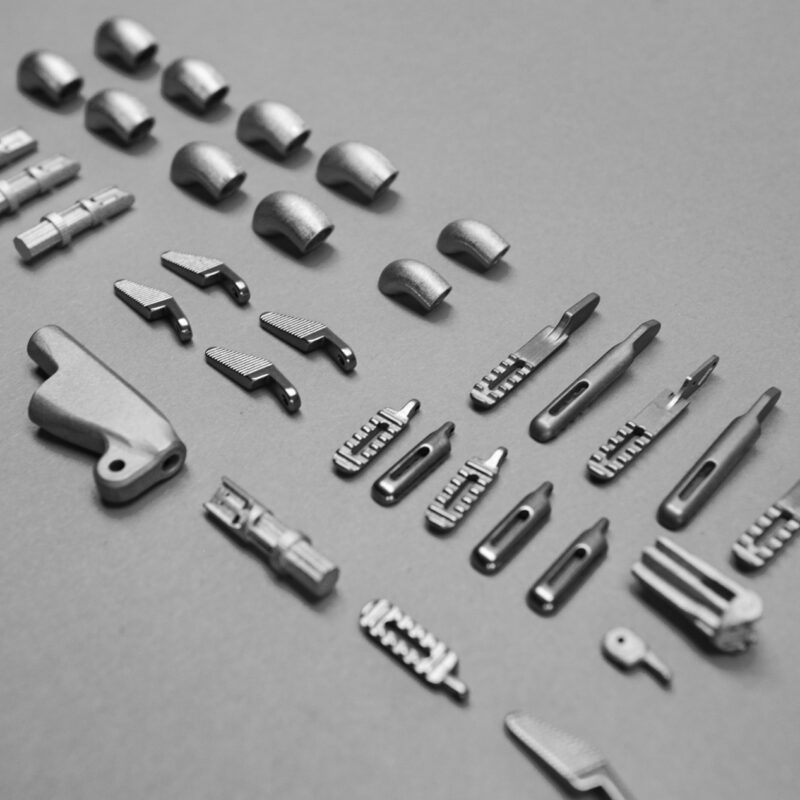 A gray surface displays an organized arrangement of various small metal components, including fittings, connectors, and hardware pieces, aligned in neat rows—an impressive showcase from a leading 3D metal printing company.