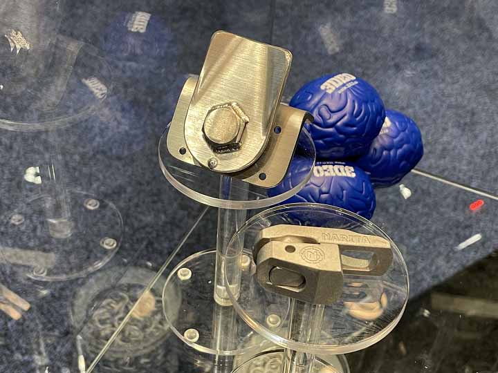 A display with two small metal mechanical components on clear stands, and multiple blue stress balls shaped like brain models in the background, highlights 3DEO talks pure copper and 3D printing for rail with 3DPrint.com.