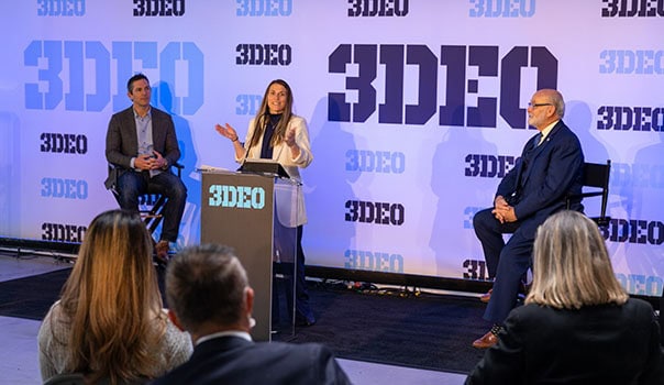 A woman is speaking at a podium labeled "3DEO" with two men seated on either side of her, discussing innovations in metal 3D printed parts. The background features the "3DEO" logo repeated multiple times. Several people are seated facing them, eagerly listening to the presentation.