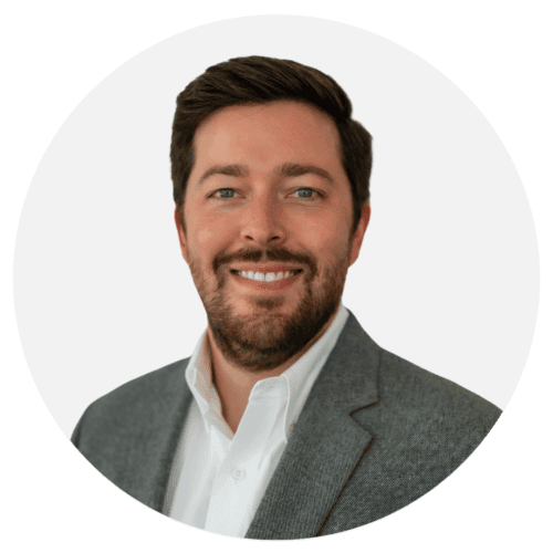 A man with dark hair and a beard smiles at the camera. He is wearing a white shirt and a gray blazer, exuding confidence much like the precision of metal 3D printed parts. The background is plain white.