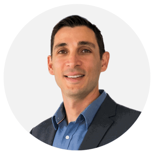 A man with short dark hair, wearing a blue shirt and a dark blazer, smiling in front of a plain white background, proudly showcasing metal 3D printed parts.