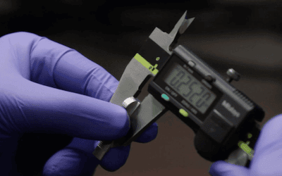 A person wearing purple gloves measures a small metal object with a digital caliper reading 0.2520 inches.