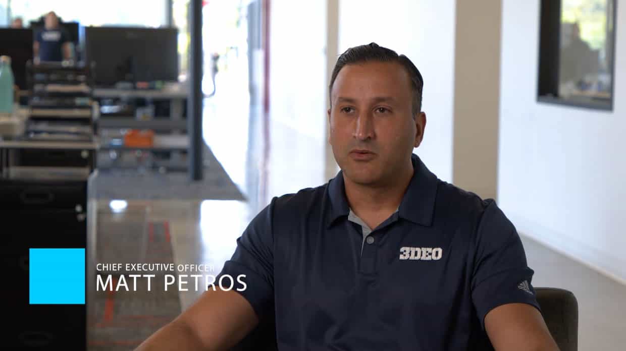 A man, identified as Chief Executive Officer Matt Petros, is seated indoors in an office setting, wearing a navy blue polo shirt with the logo "3DEO," a company renowned for its innovative metal 3D printed parts.